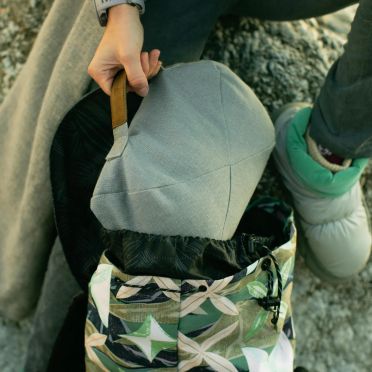 Zbliżenie na osobę wyciągającą jasnoszarą poduszkę do medytacji z kolorowego plecaka, w tle kamienne podłoże i zimowy strój sugerujący aktywność na świeżym powietrzu.